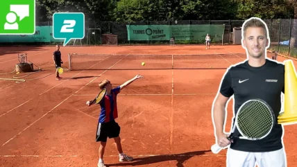 Young boy serving on a clay tennis court while a coach or pro watches; large cutout of a man with a racket on the right; banners in the background