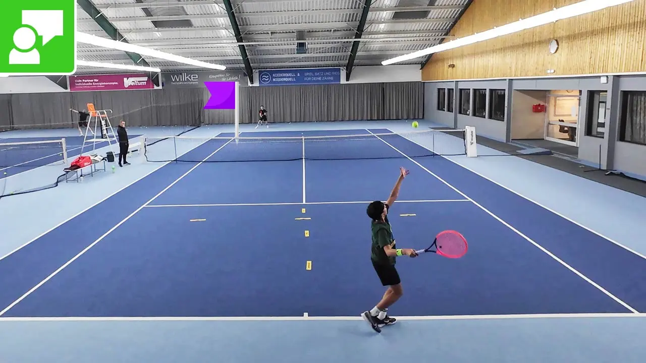 Indoor tennis court with a player serving near the baseline; blue flooring, net, referee chair on the left, and another player in the back.