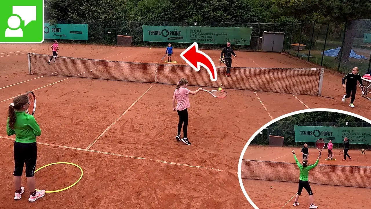 Children practice tennis on a red clay court during a drill; a girl serves while others wait near the net, with a coach nearby and banners in the background.